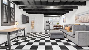Ein modernes Wohnzimmer mit einem schwarz-weißen Schachbrettboden, einem grauen Sofa, einem Holzesstisch und einer Küche im Hintergrund.