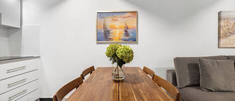 Un coin repas avec une table en bois, des chaises, un vase de fleurs, un tableau représentant un coucher de soleil et un canapé gris.