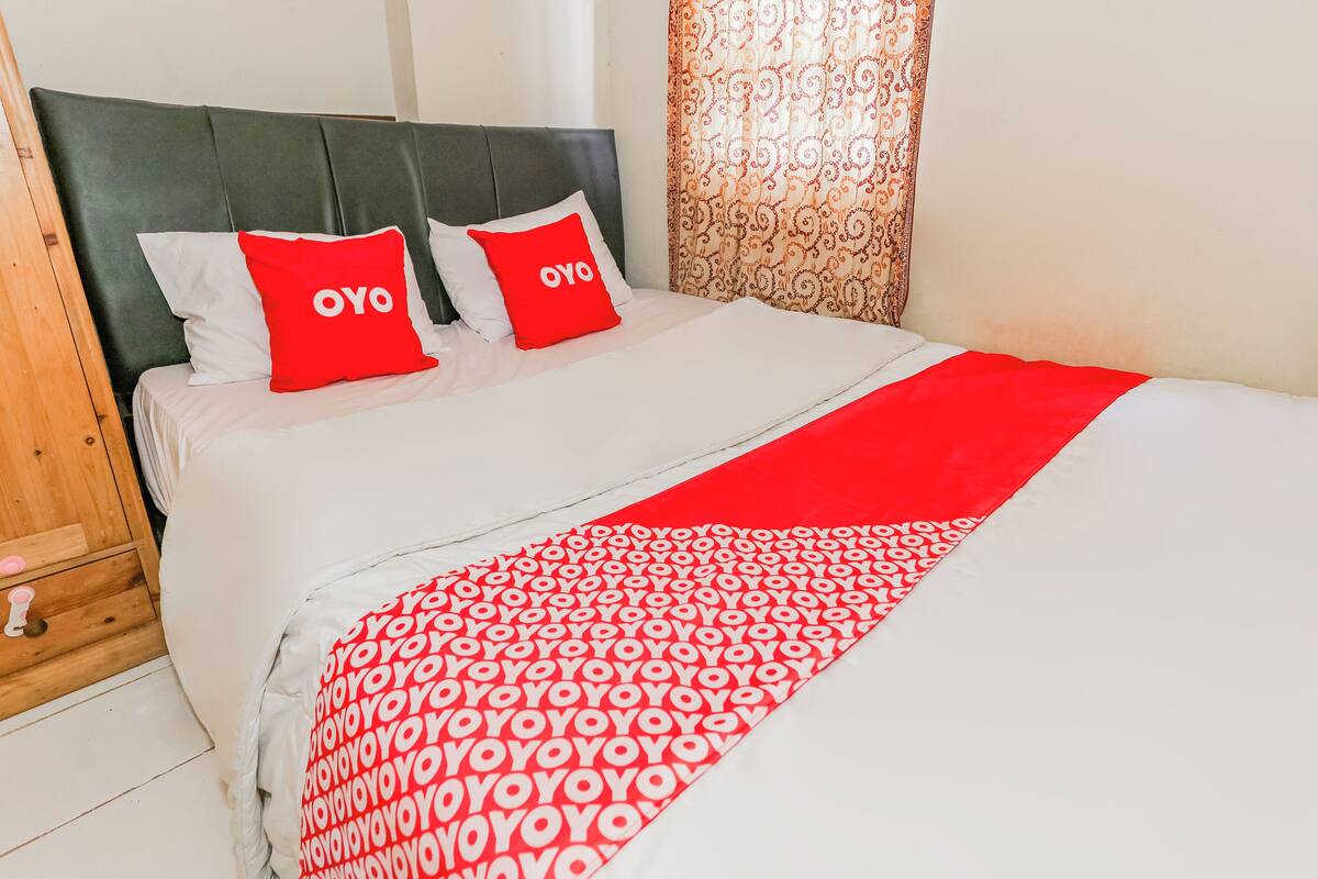 A neatly made bed with a white and red bedspread, two pillows with the word "OYO" on them, and a wooden nightstand with a drawer.