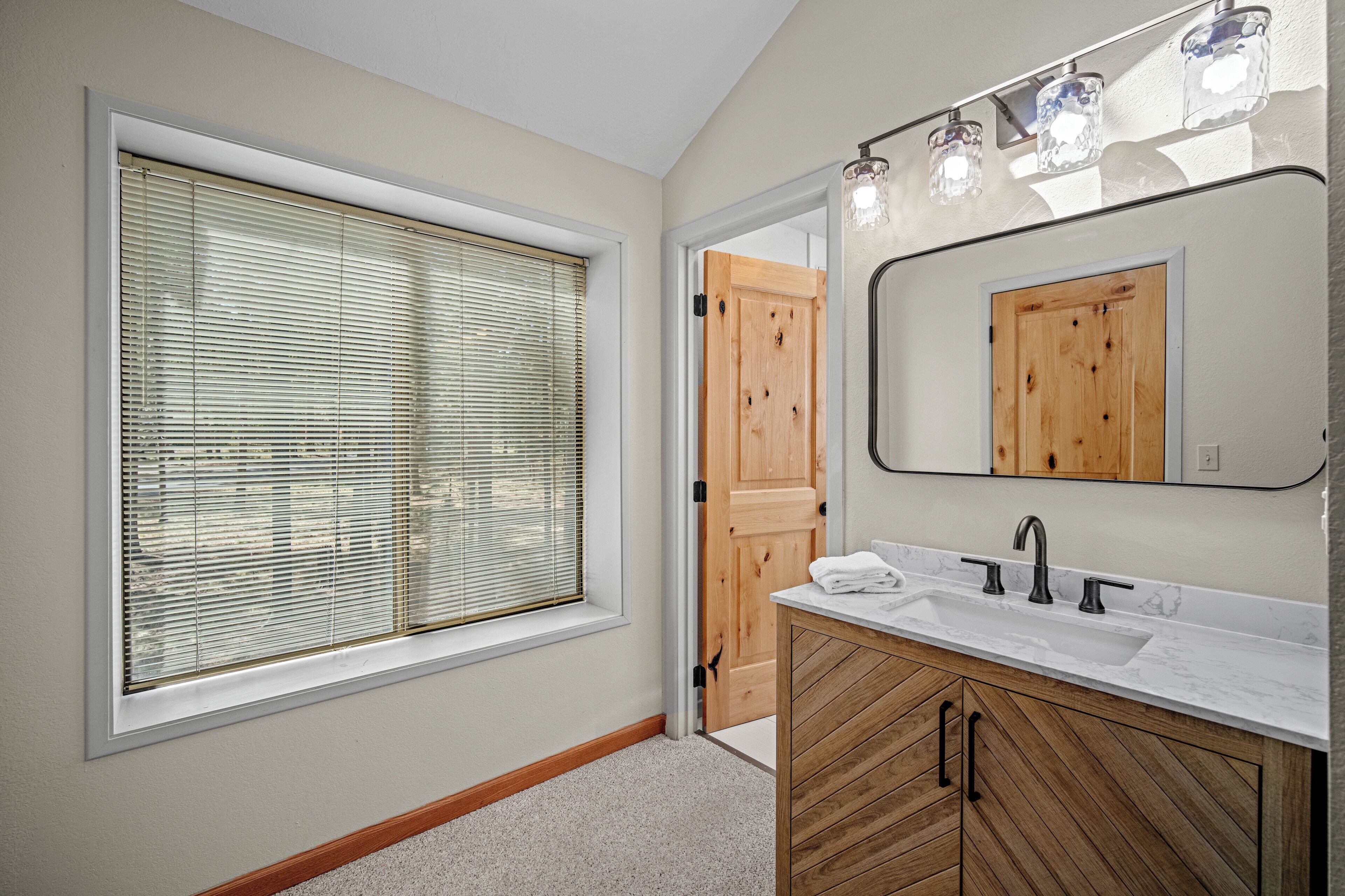 A bathroom with a large window, wooden vanity, and a mirror with lights.