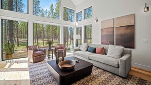 A spacious living room with large windows, a gray sofa, a brown leather chair, and a wooden coffee table. - Family-Friendly Caldera Retreat with Access to Exclusive Golf (Bend)