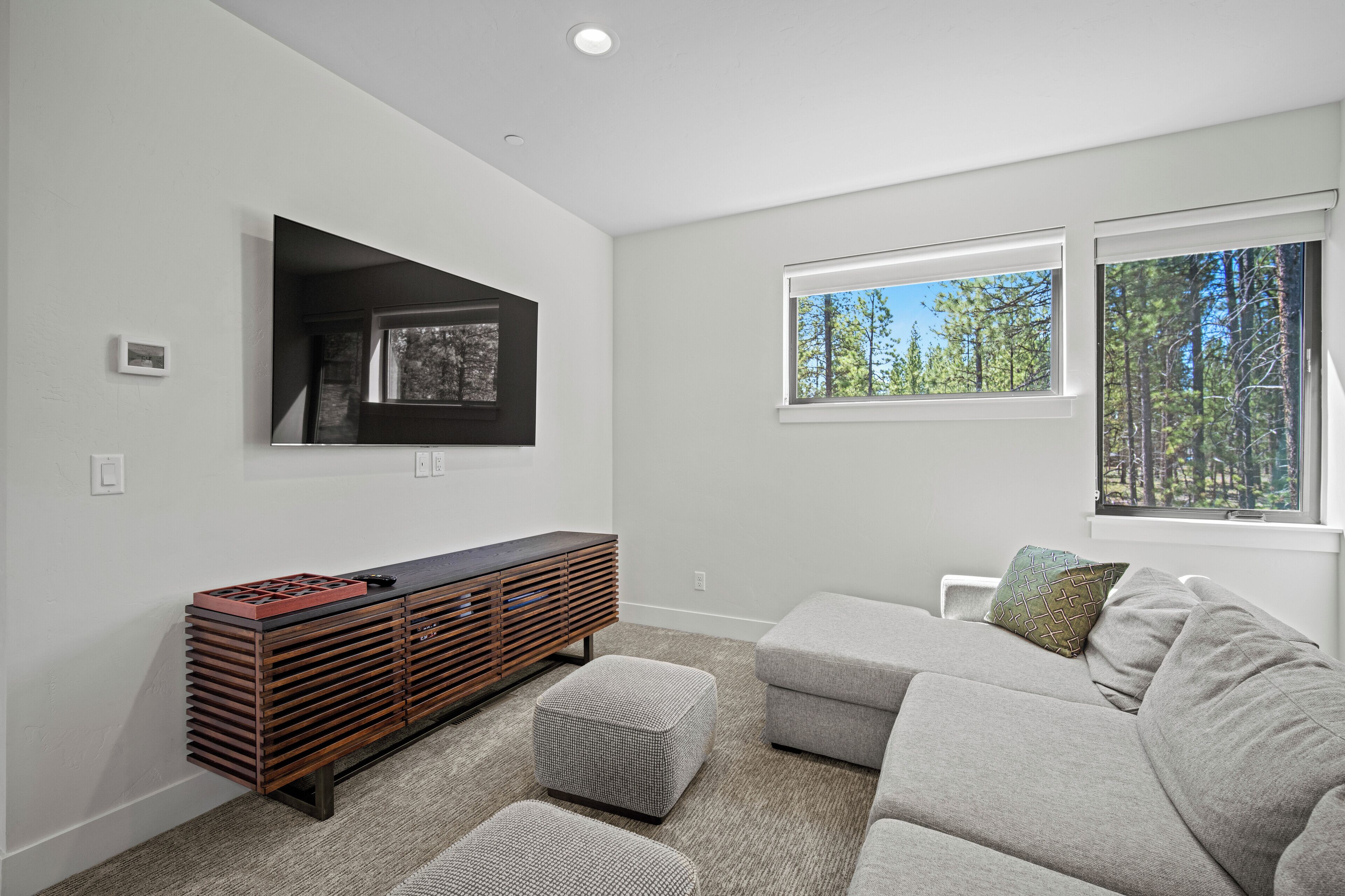 A modern living room with a sectional sofa, a flat-screen TV mounted on the wall, a wooden entertainment unit, and large windows offering a view of trees.