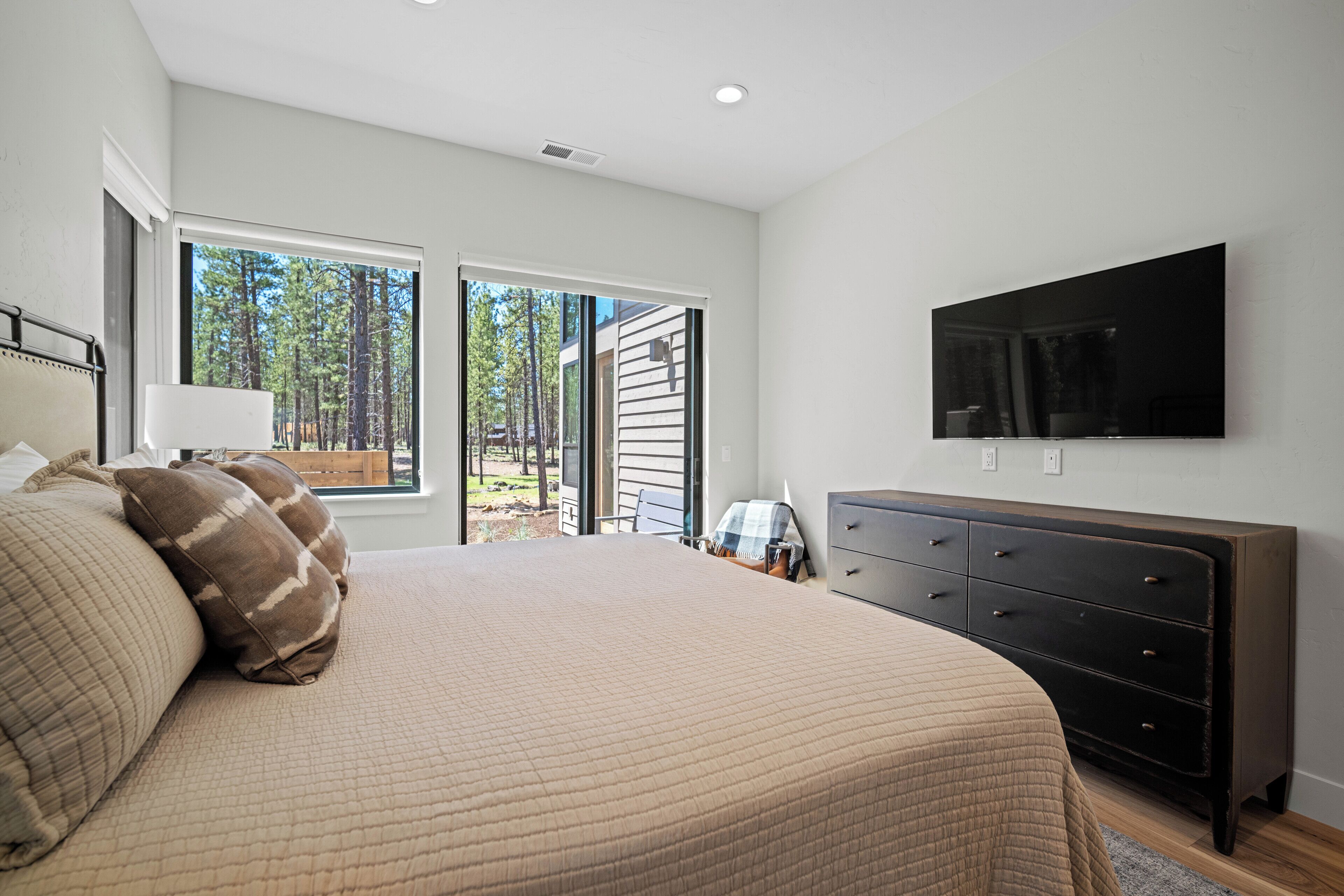 A bedroom with a large bed, a dresser, a TV mounted on the wall, and a sliding glass door leading to a balcony with a view of trees.