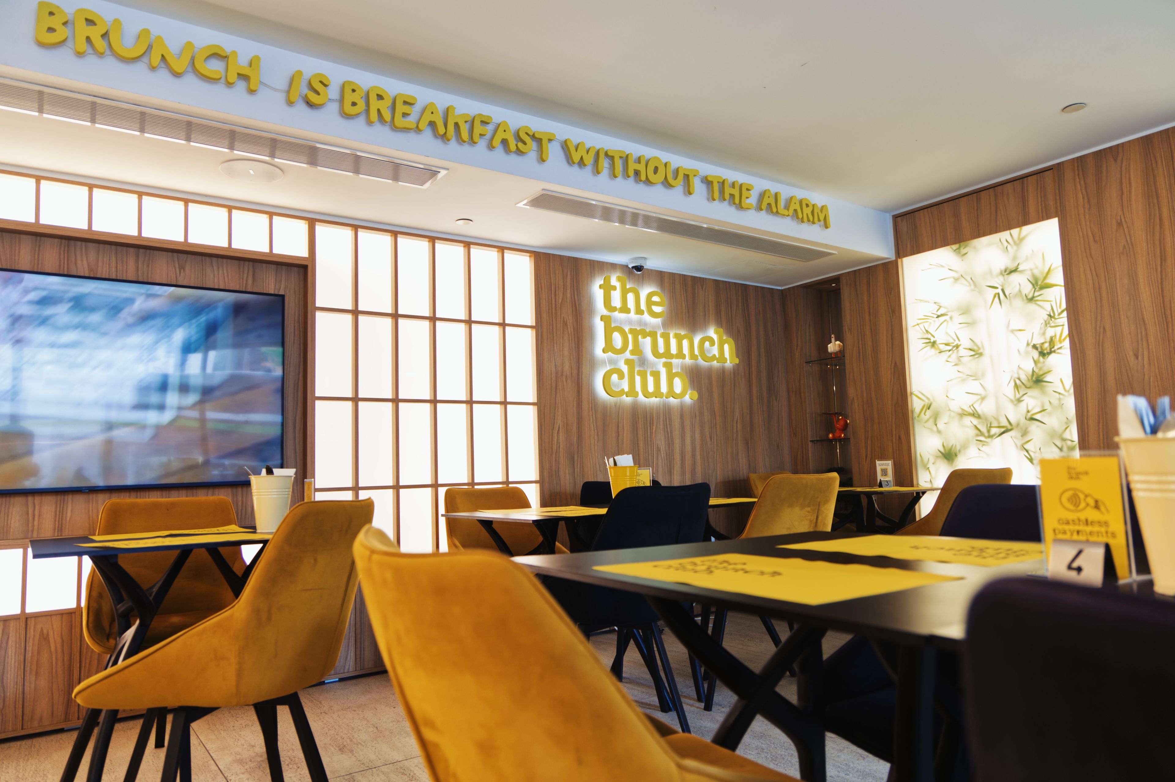 Un espace repas moderne avec un grand écran de télévision, une pancarte indiquant « LE BRUNCH, C’EST LE PETIT-DÉJEUNER SANS LE RÉVEIL », et une autre pancarte pour « le club du brunch ».