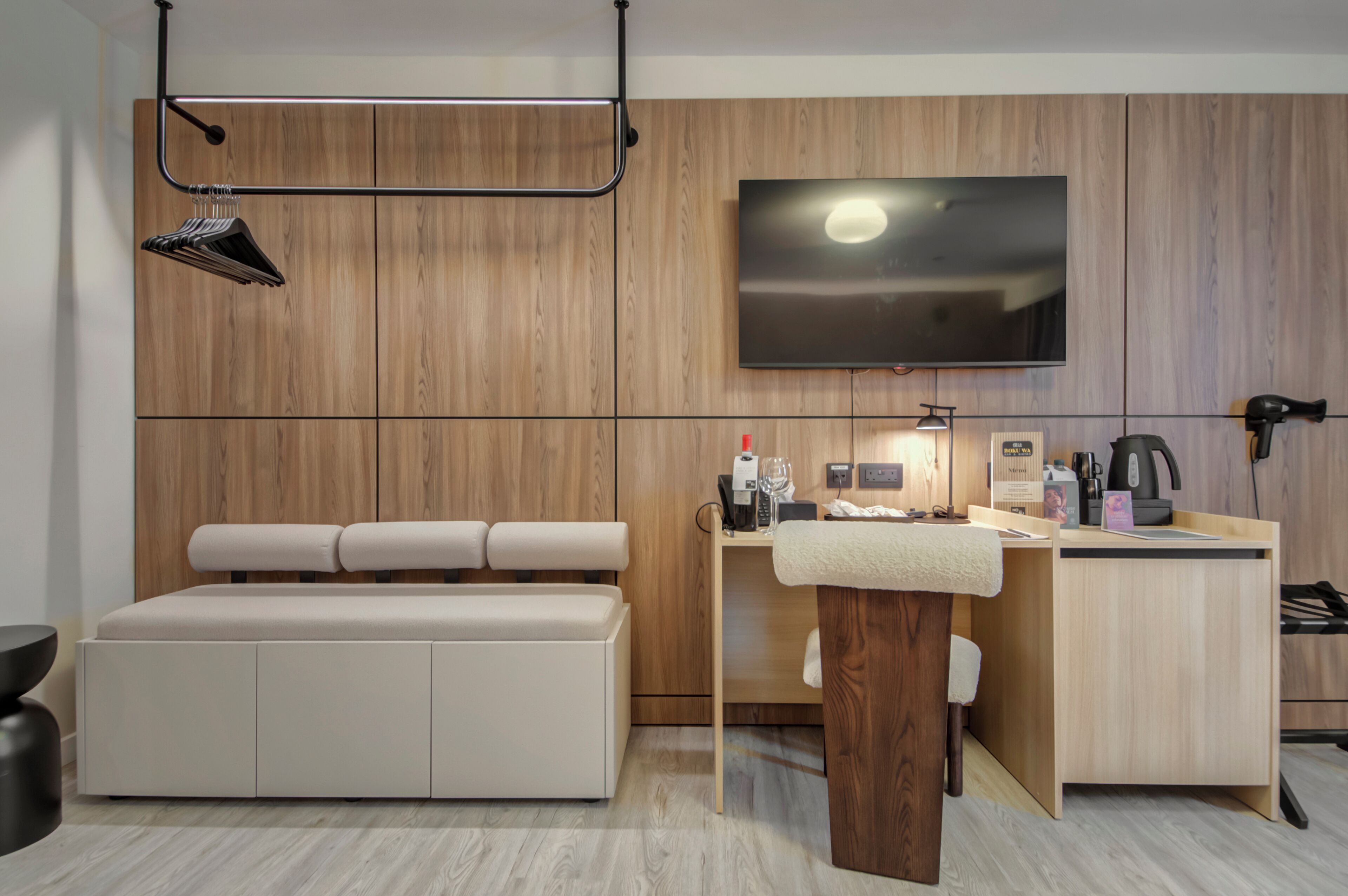 A modern hotel room with a wooden panel wall, a flat-screen TV, a sofa, a small desk with a coffee maker, and a hairdryer.