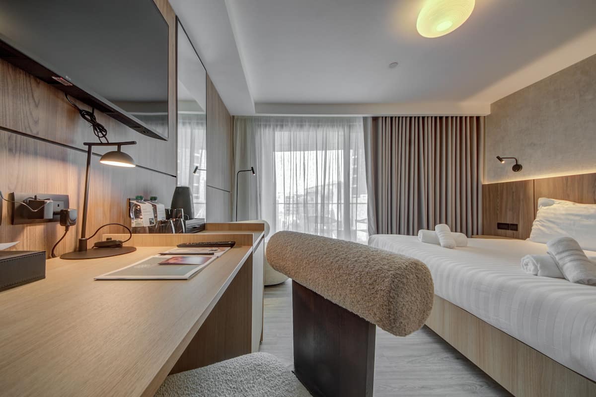 A modern hotel room with a wooden desk, a bench, a bed with white linens, and a large window with curtains.