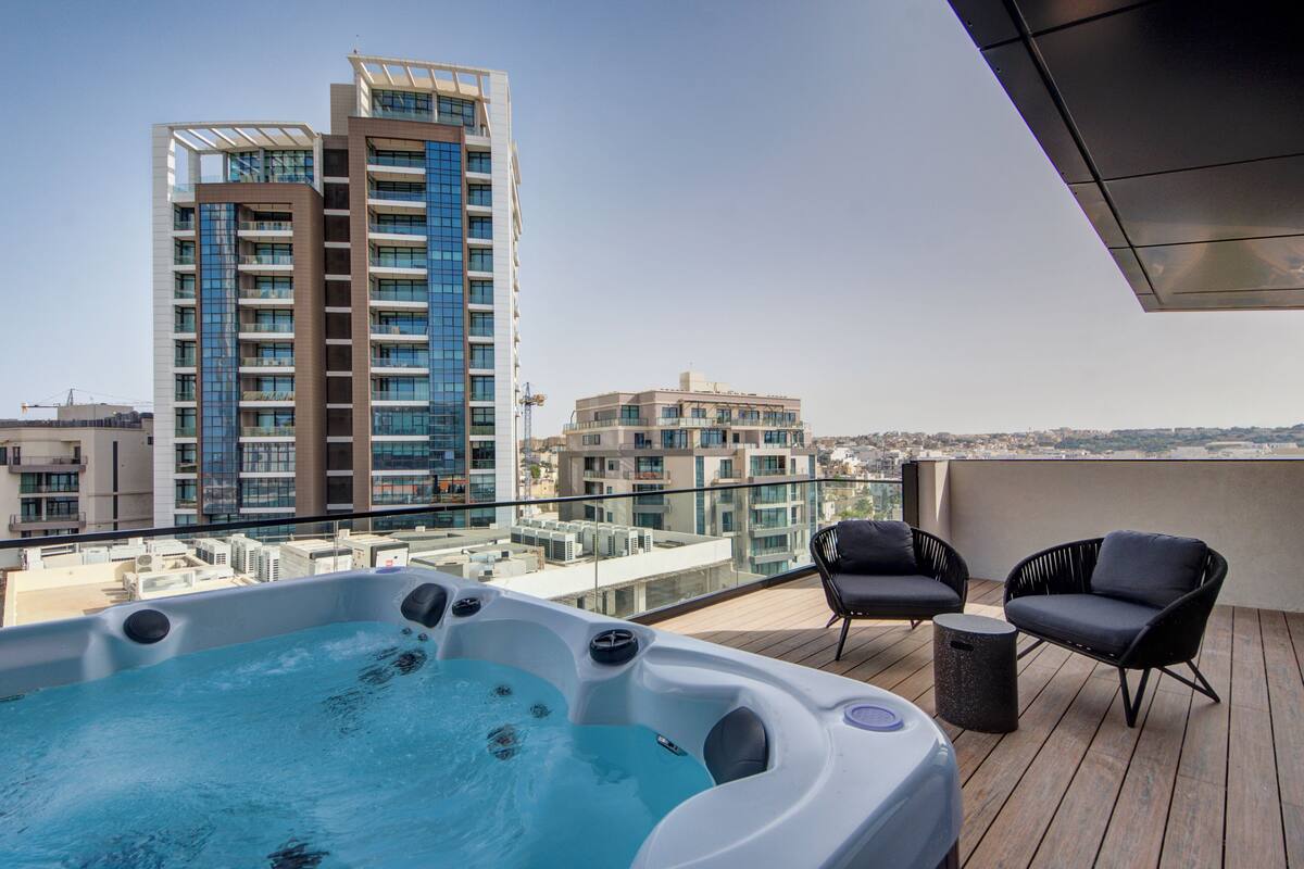 A rooftop terrace with a hot tub, two black wicker chairs, and a view of high-rise buildings.