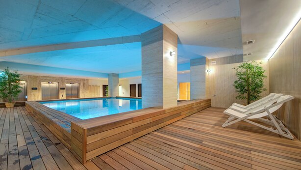 Una piscina interior con plataforma de madera y sillas de descanso, rodeada de paredes con iluminación empotrada.