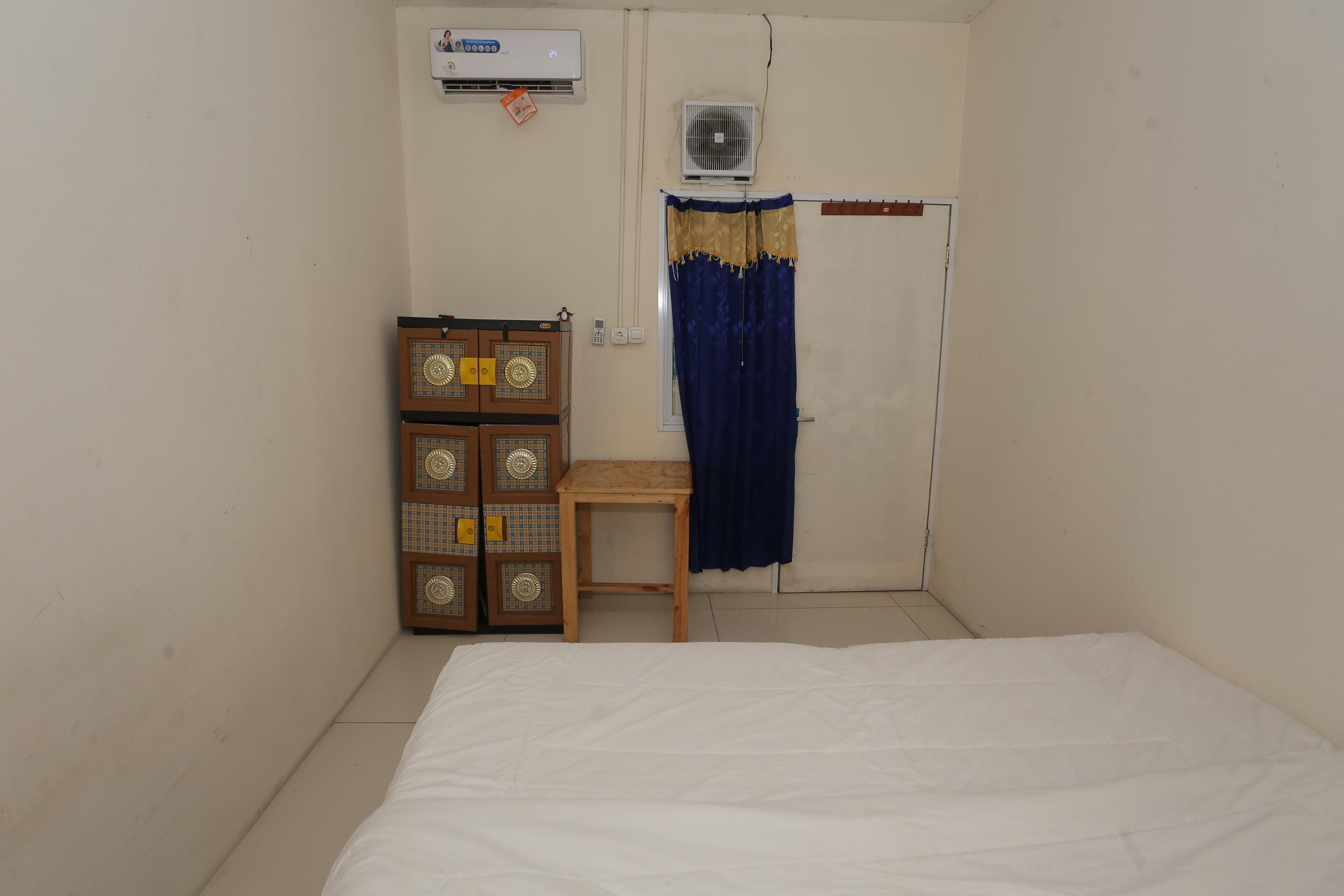 A small, clean room with a bed, a wooden cabinet, a small table, a door with a curtain, and an air conditioning unit.