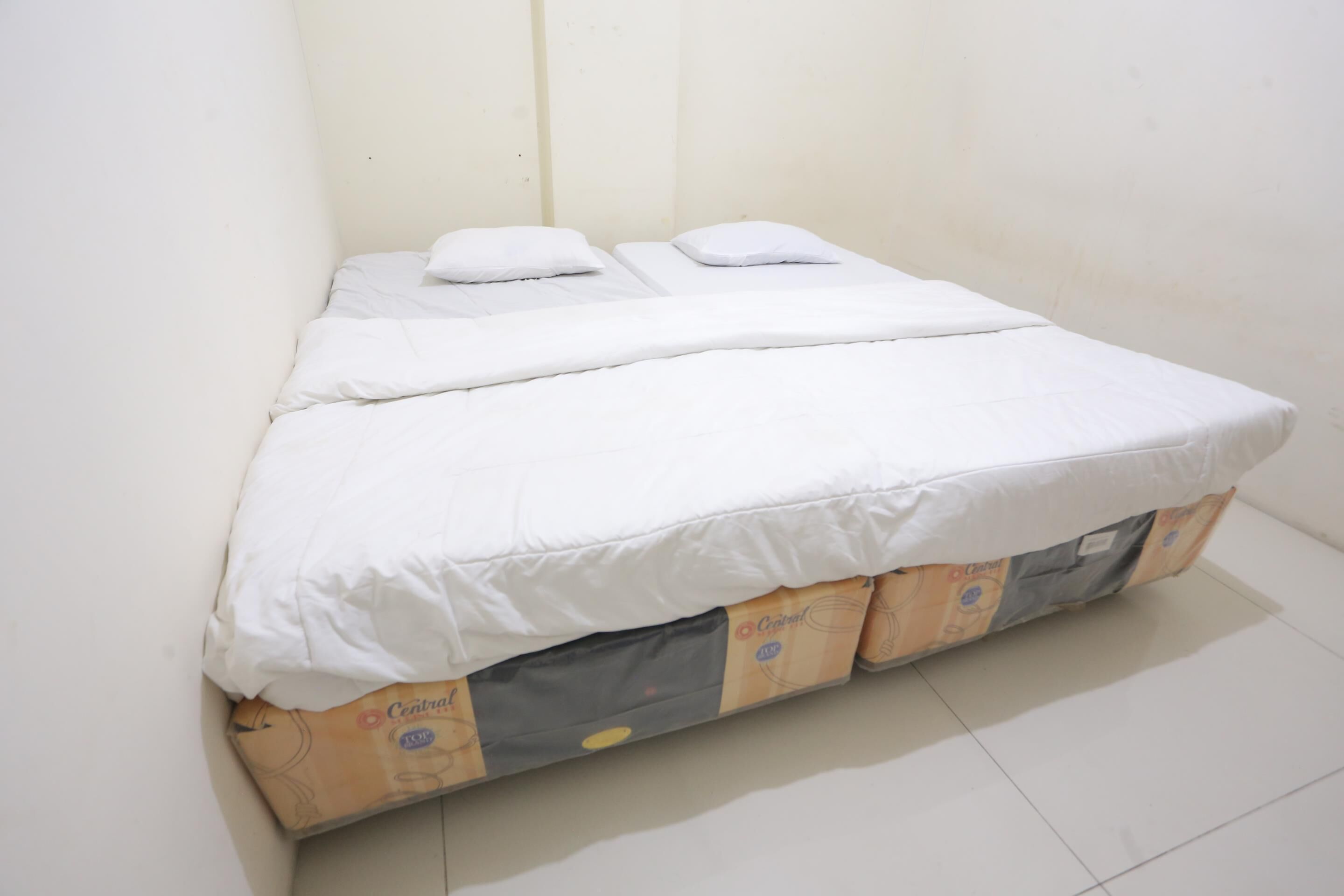 A small, single-bed room with a white mattress and pillows. The bed is placed against a plain wall, and there is a visible mattress protector underneath. The room has a tiled floor and a simple, functional design.