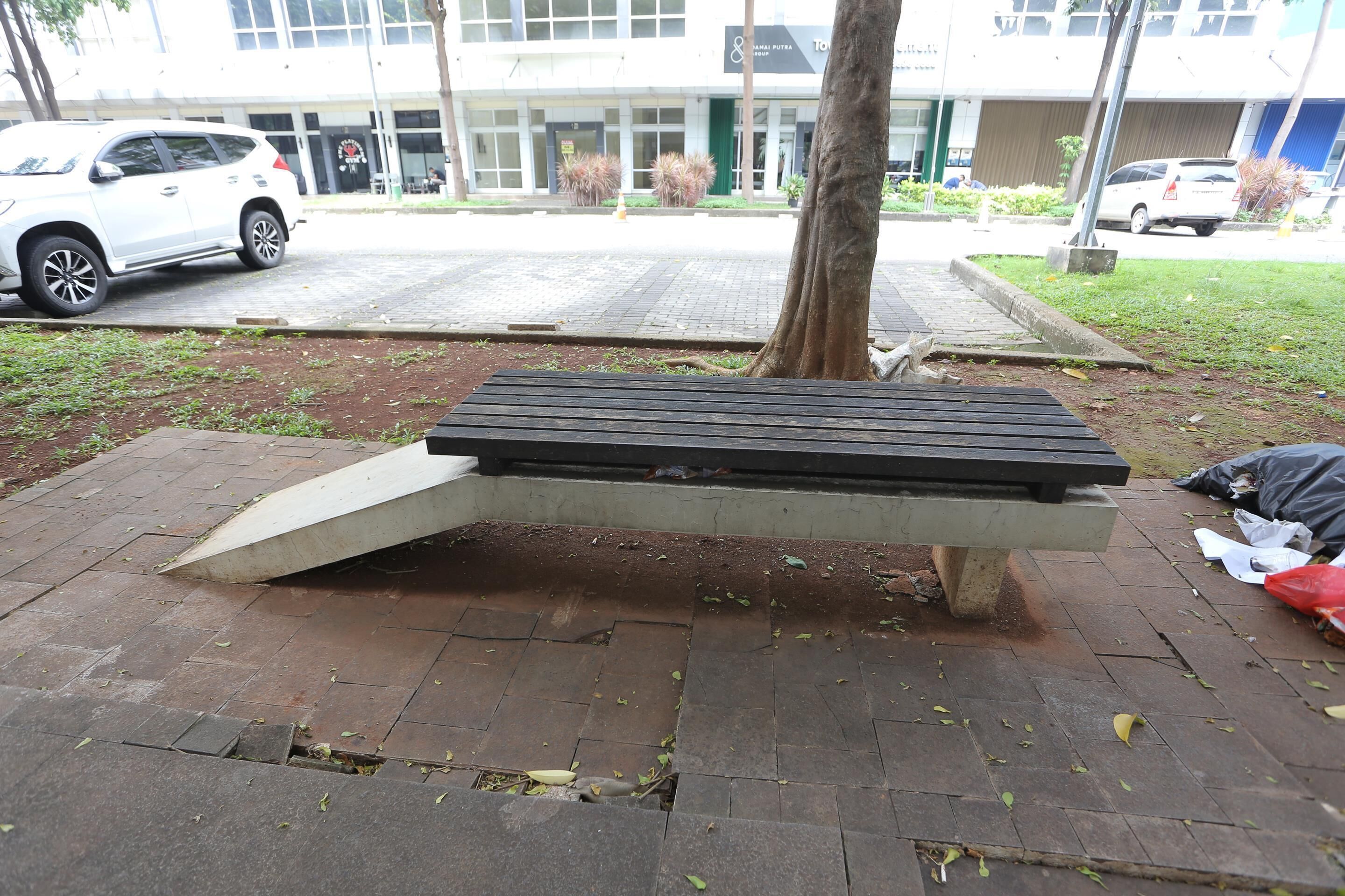 A park bench with a slanted backrest, surrounded by a paved area with scattered leaves.