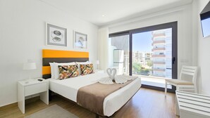 A modern hotel room with a bed, a nightstand, a desk, a chair, and a large window offering a city view.