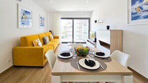 A modern living room with a yellow sofa, a dining table set for two, and a kitchen area visible through an open door. - Flat Praia Rocha Residence Honey Glow (Portimão)