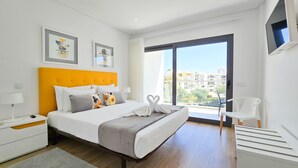 A modern bedroom with a large bed, a bedside table, a chair, and a view of the city through a large window. - Flat Praia Rocha Residence Honey Glow (Portimão)