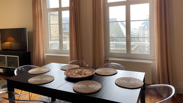A dining area with a table set for four, a television, and large windows with curtains. - 2 Bedroom Apartment in Saint-Malo (Saint-Malo)