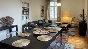 A modern living room with a dark dining table, clear chairs, and woven placemats. There is a bookshelf, a sofa, and a lamp in the room. - 2 Bedroom Apartment in Saint-Malo (Saint-Malo)