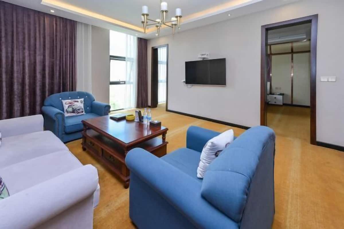 Ein modernes Wohnzimmer mit einem blauen Sofa, einem weißen Sessel und einem Holztisch. Ein Flachbildfernseher ist an der Wand angebracht, und es gibt eine Schiebetür zu einem anderen Raum.