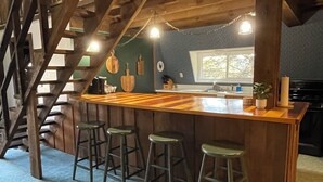 A rustic bar area with wooden beams, a staircase, and a kitchen in the background. - Big Barn in Bemus Point, unique, peaceful, and memorable (Bemus Point)