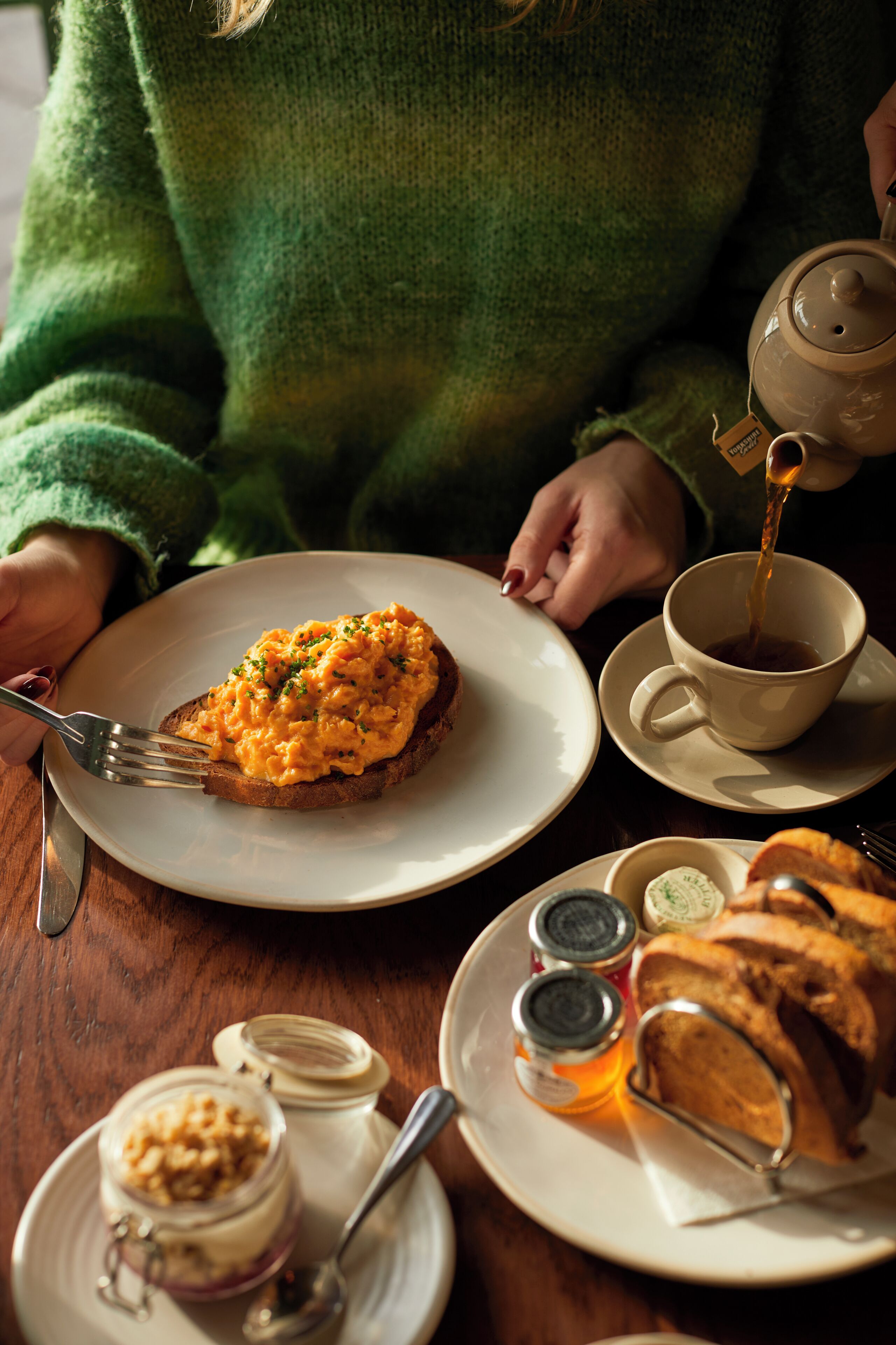 A dining table with a plate of scrambled eggs, a cup of tea, and a jar of jam.