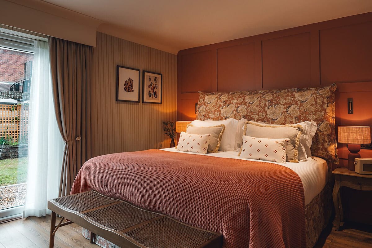 Un dormitorio con cama, mesita de noche, lámpara, ventana con cortinas y vistas al exterior.