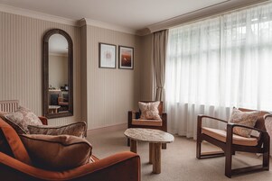 Una habitación con papel tapiz a rayas, un espejo grande, dos cuadros enmarcados, una mesa redonda y tres sillas de madera con cojines.