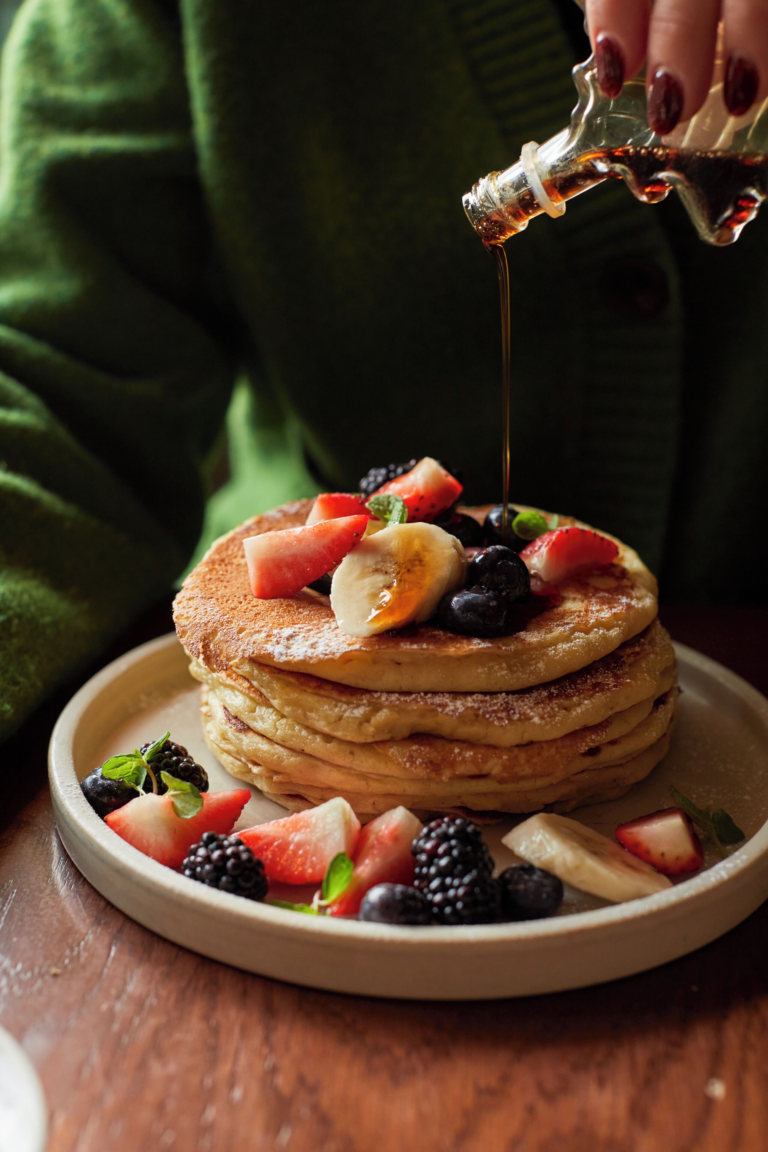 A stack of pancakes topped with various fruits and syrup being poured on them.