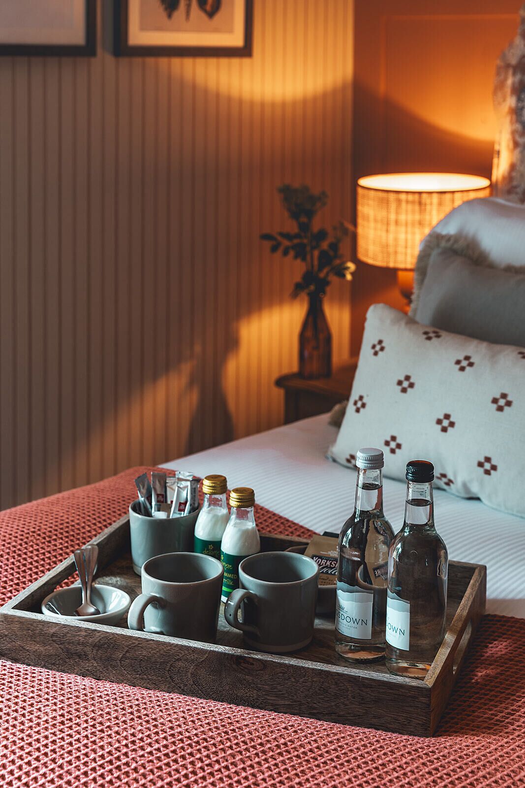 A tray with various bottles and mugs on a bed with a patterned pillow and a lamp in the background.