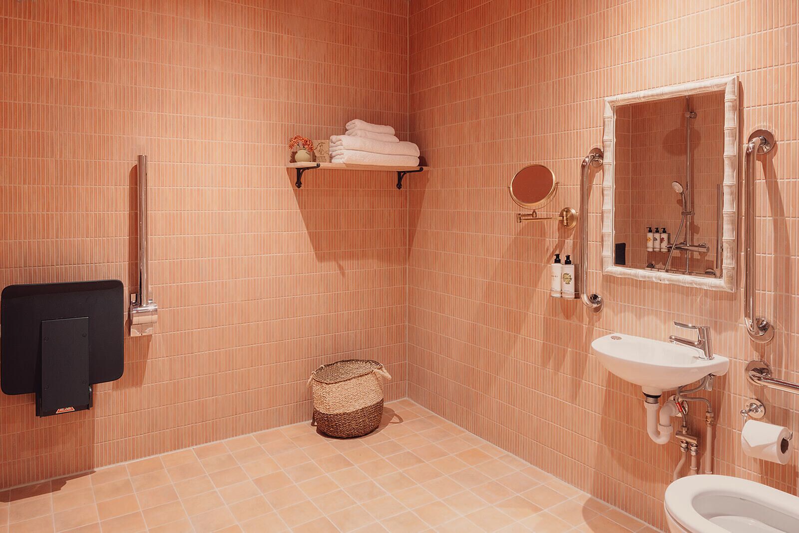 Baño con inodoro, lavamanos y ducha. Hay un perchero, una cesta y una estantería con artículos de aseo.