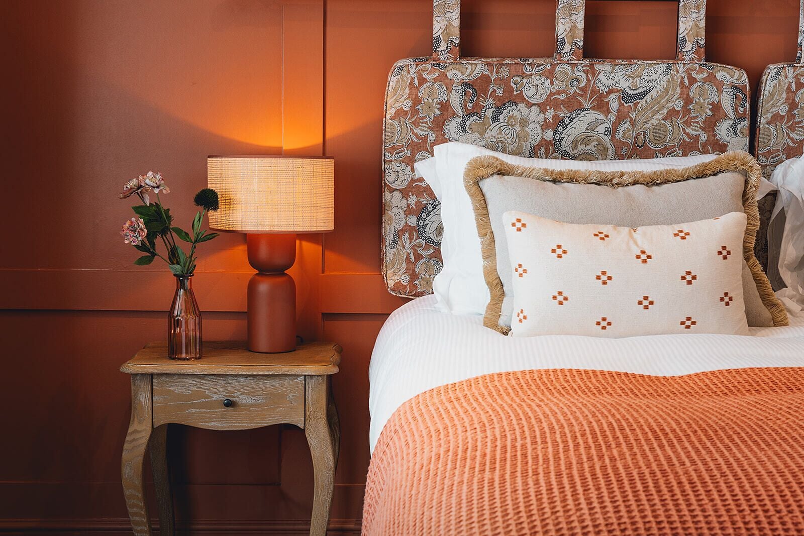 Un dormitorio con un cabecero estampado, una mesita de noche con lámpara y un arreglo floral.