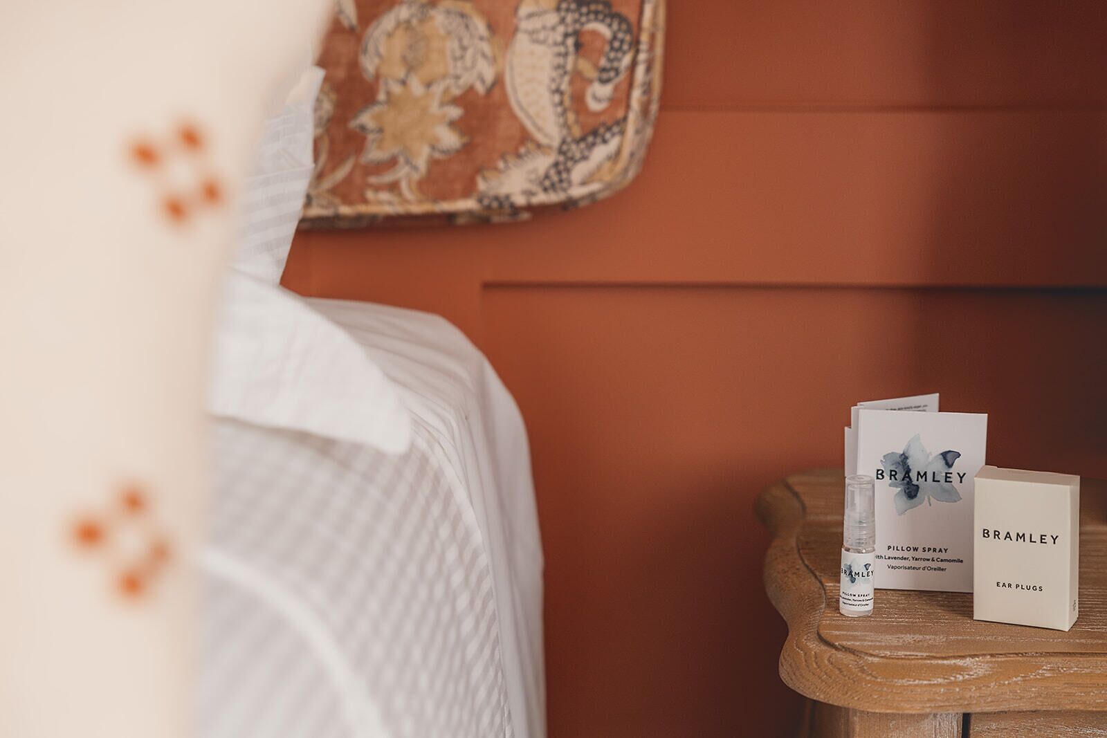 Een net opgemaakt bed met een witte spreideken, een klein houten nachtkastje met Bramble Pillow Spray en een doosje met 'Oordoppen'.