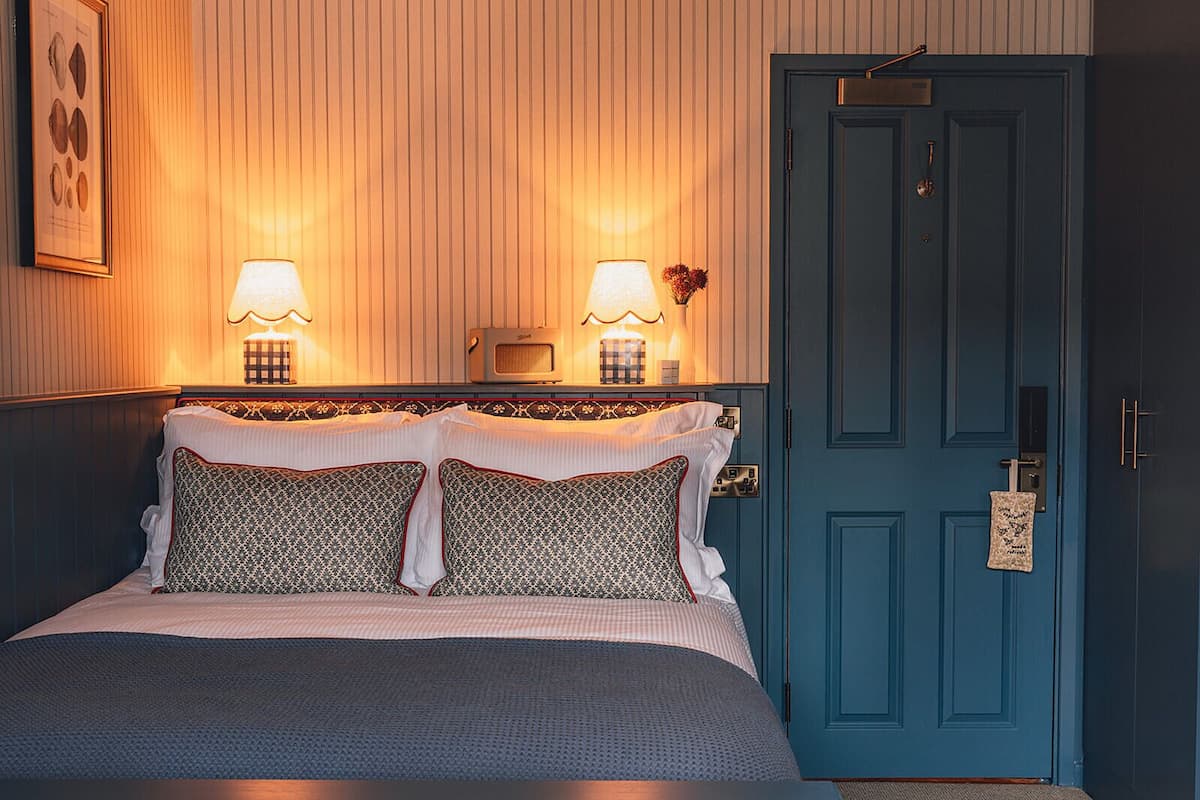 Una cama bien hecha con almohadones estampados, una puerta azul con cerradura y dos lámparas fijadas a la pared.