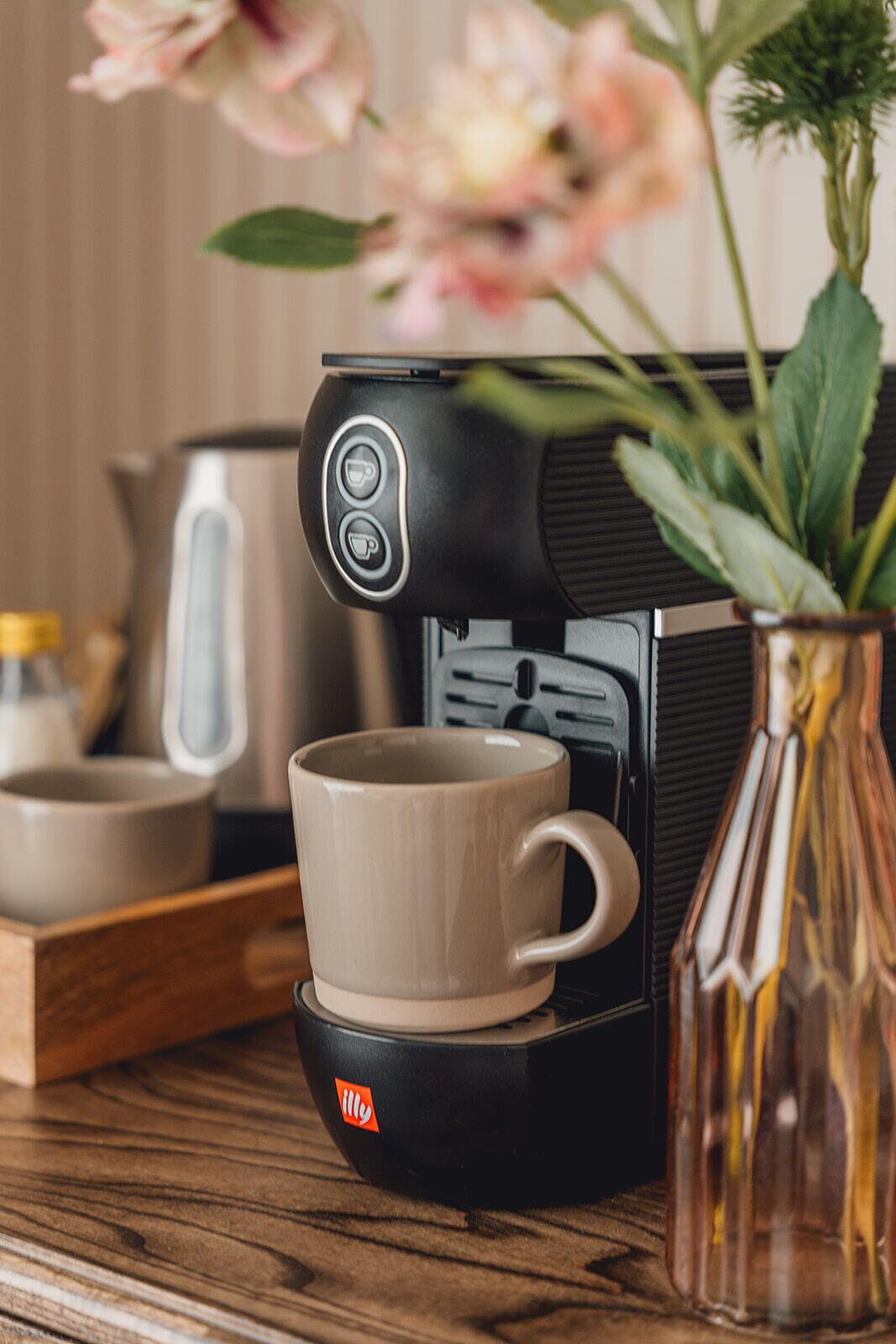A coffee machine with a cup below it, a vase with flowers, and a kettle in the background.