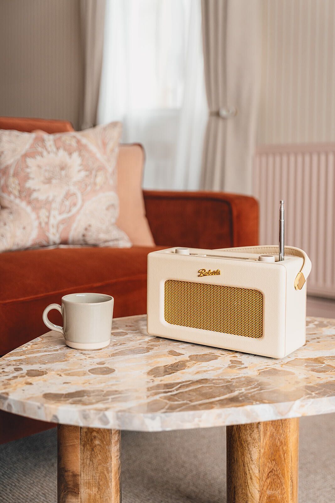 A vintage radio on a marble table with a white mug beside it, in a room with a red sofa and patterned cushions.
