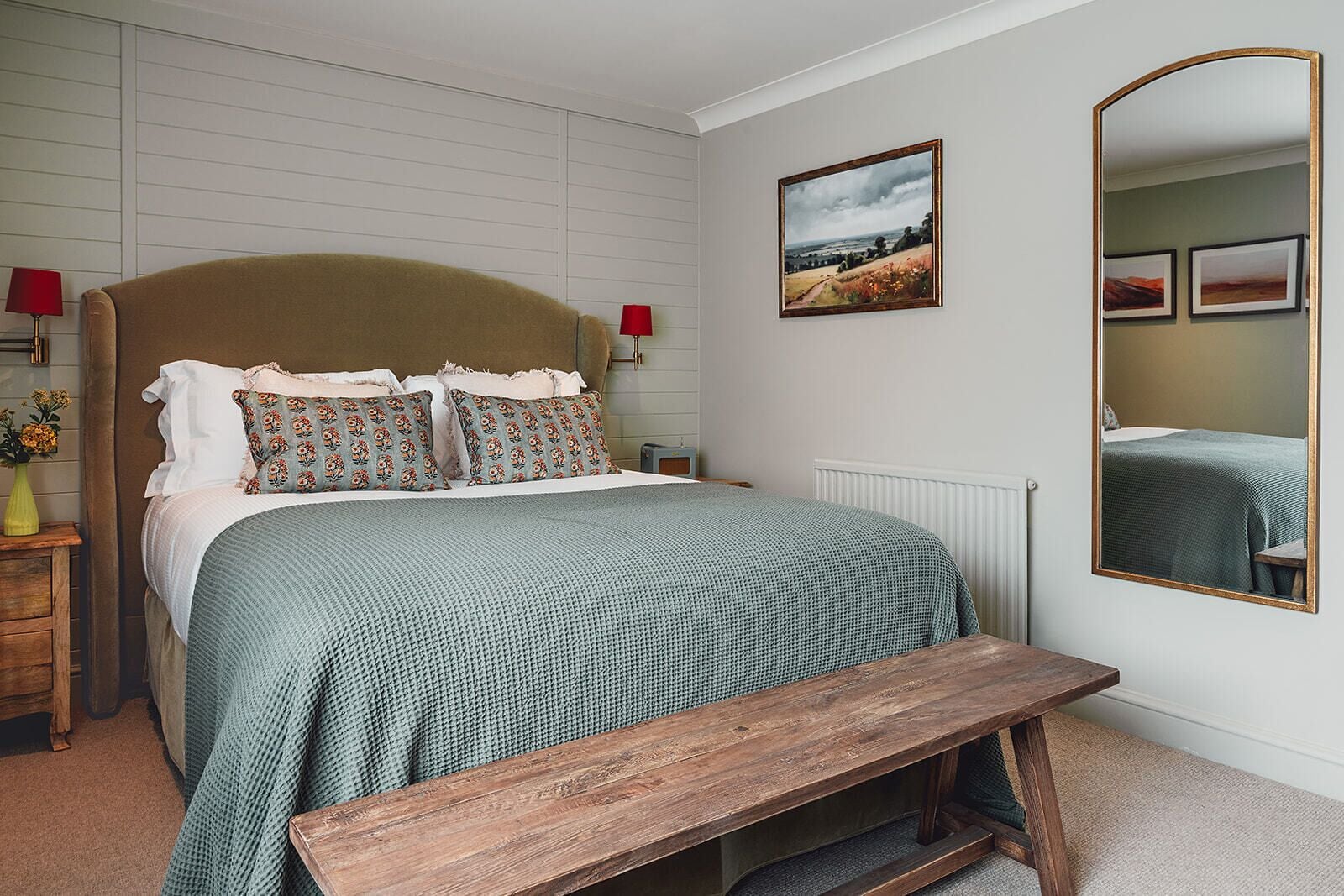 Une chambre à coucher avec un lit, un banc, un miroir et un tableau accroché au mur.