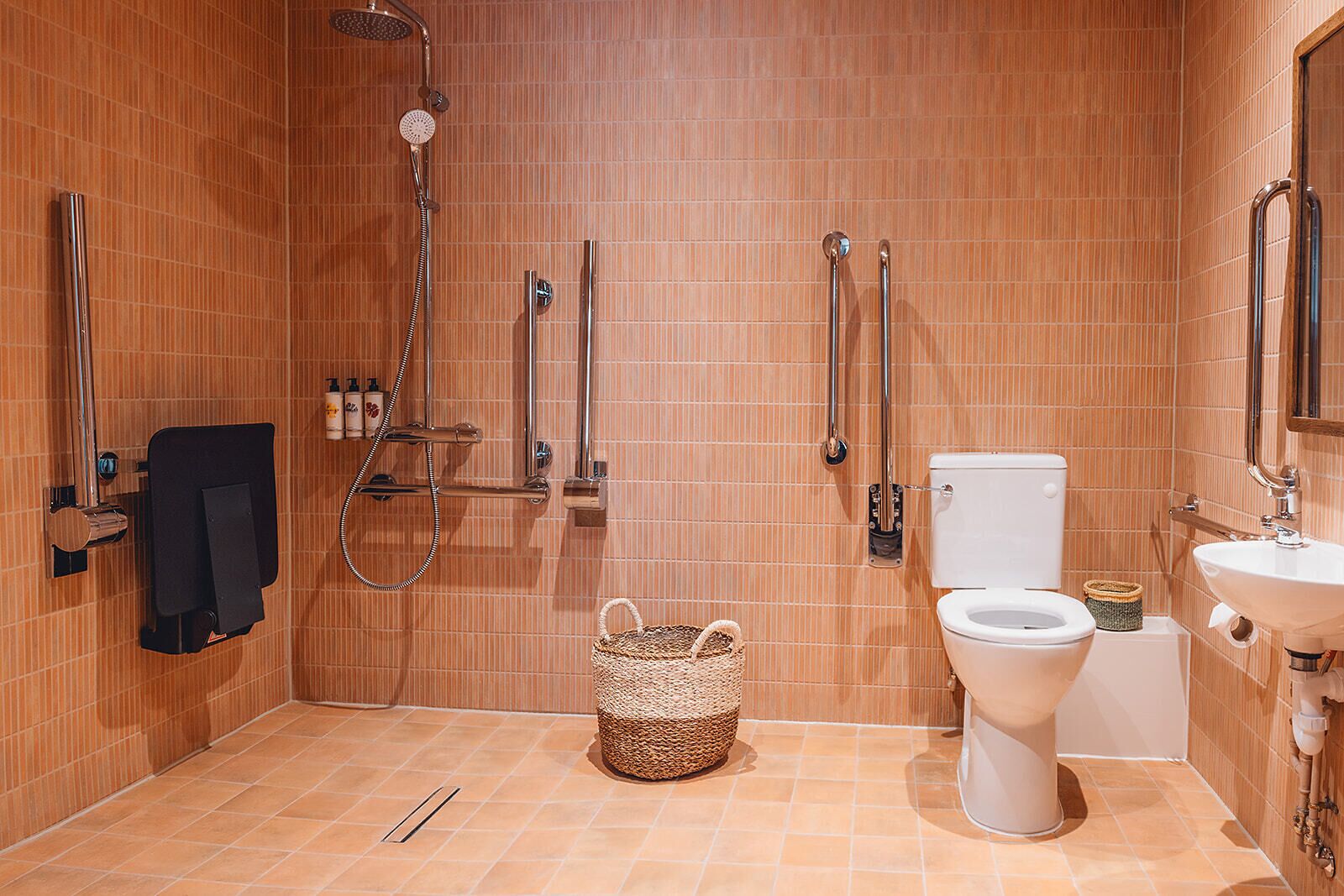 Un baño con ducha, inodoro y lavamanos, con azulejos en tonos cálidos, un perchero y una cesta en el suelo.