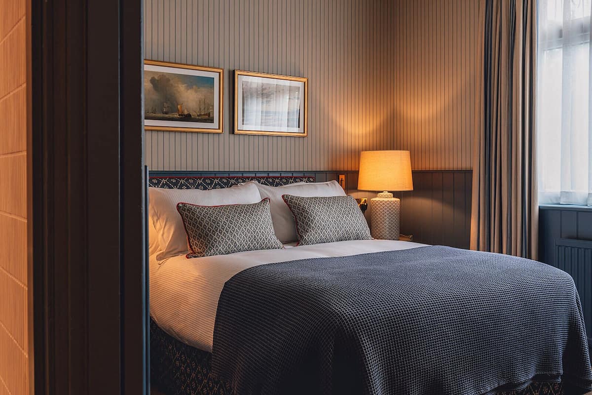 Una cama bien hecha con almohadones estampados, una lámpara de noche y dos cuadros enmarcados en la pared.