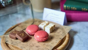 A tray with macarons and chocolates on a wooden board, a coffee cup, and books in the background.