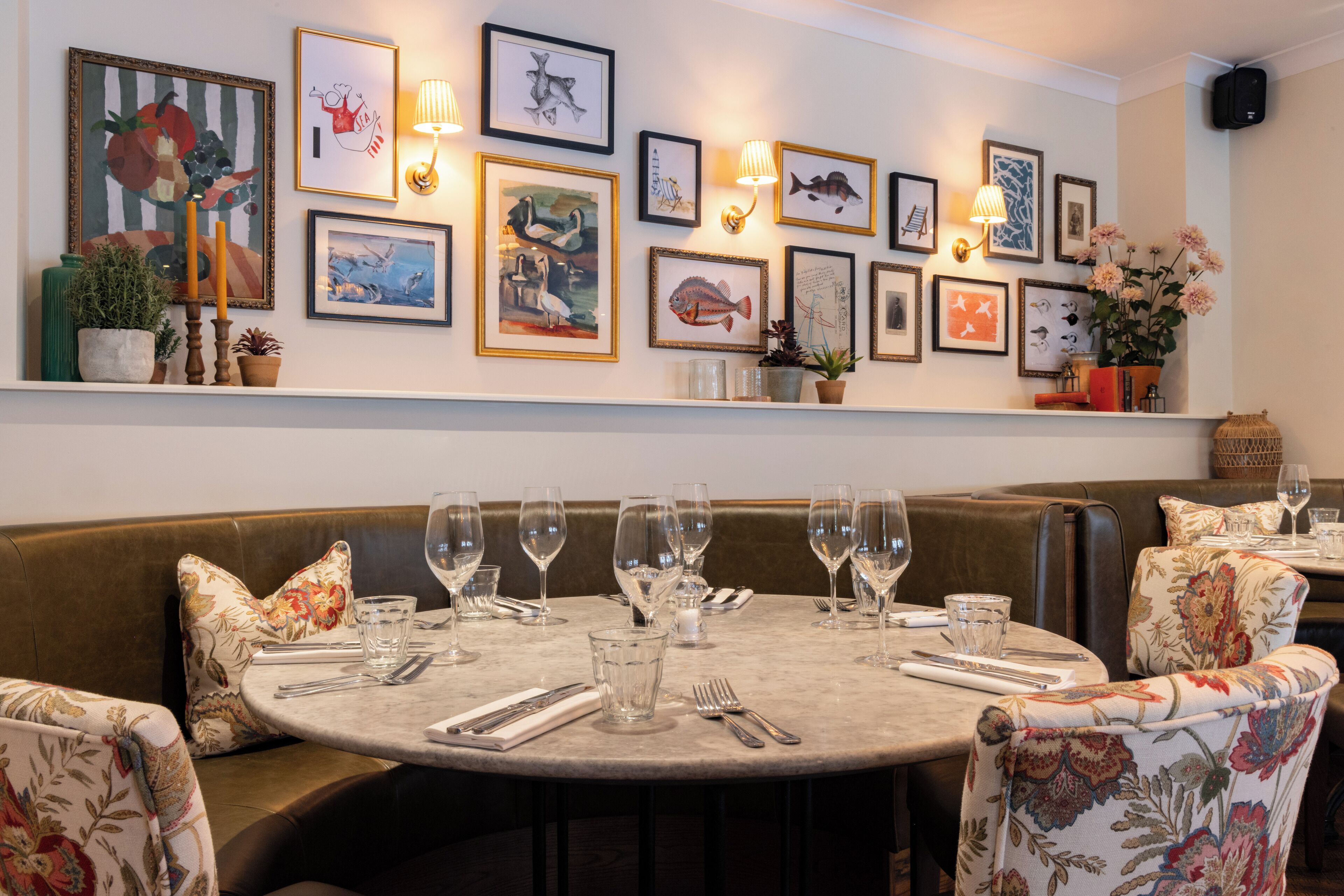 A dining area with round tables set for service, a variety of framed artworks on the wall, and a selection of plants and candles on the shelves.