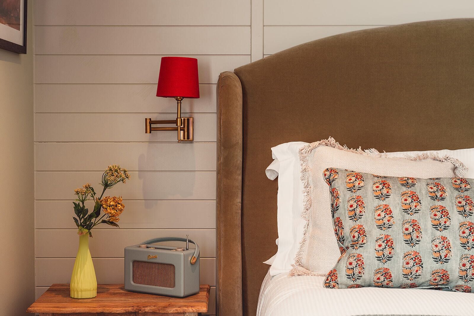 Une chambre à coucher avec une tête de lit marron, un oreiller blanc orné d’un motif floral, une lampe rouge, un vase jaune avec des fleurs et une radio ancienne posée sur une table de chevet en bois.