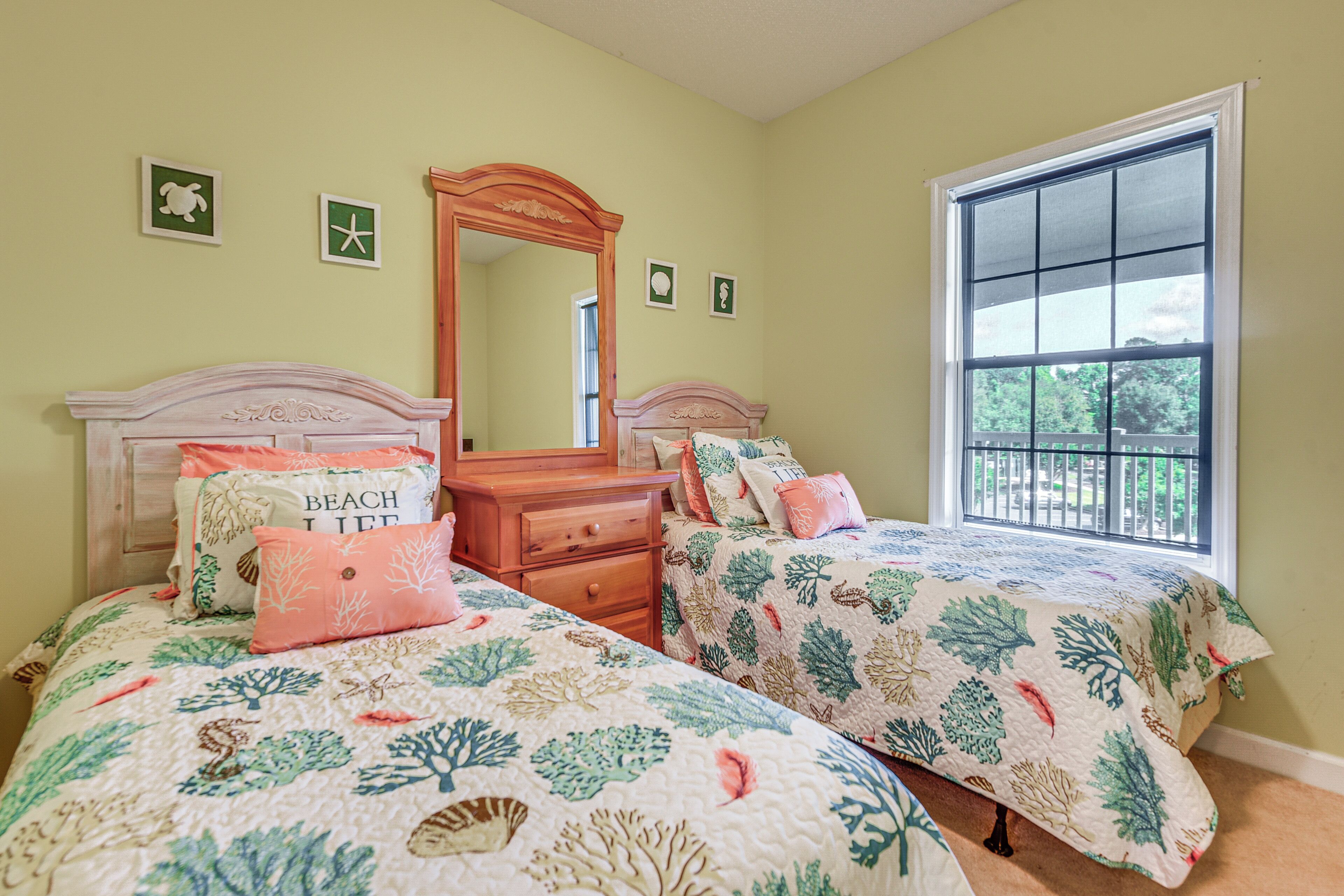 A bedroom with two beds, a dresser, a mirror, and a window with a view of greenery.