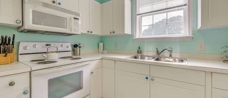 Una cocina con armarios blancos, estufa, microondas y ventana con persianas.