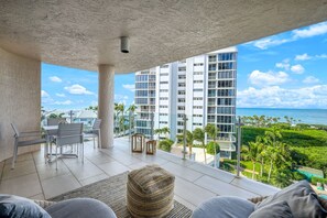 A balcony with a view of a modern building, a dining area with chairs and a table, and a woven pouf.