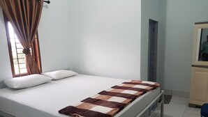 Una cama individual con colchón blanco, manta cuadricada marrón y beige, y dos almohadas blancas. Se ve una ventana con cortinas marrones, y un espejo colgado en la pared.