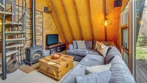 A cozy loft bedroom with a built-in fireplace, a large gray sofa, and a spiral staircase. - Cle Elum A-frame - Direct Yakima River Access! (Cle Elum)