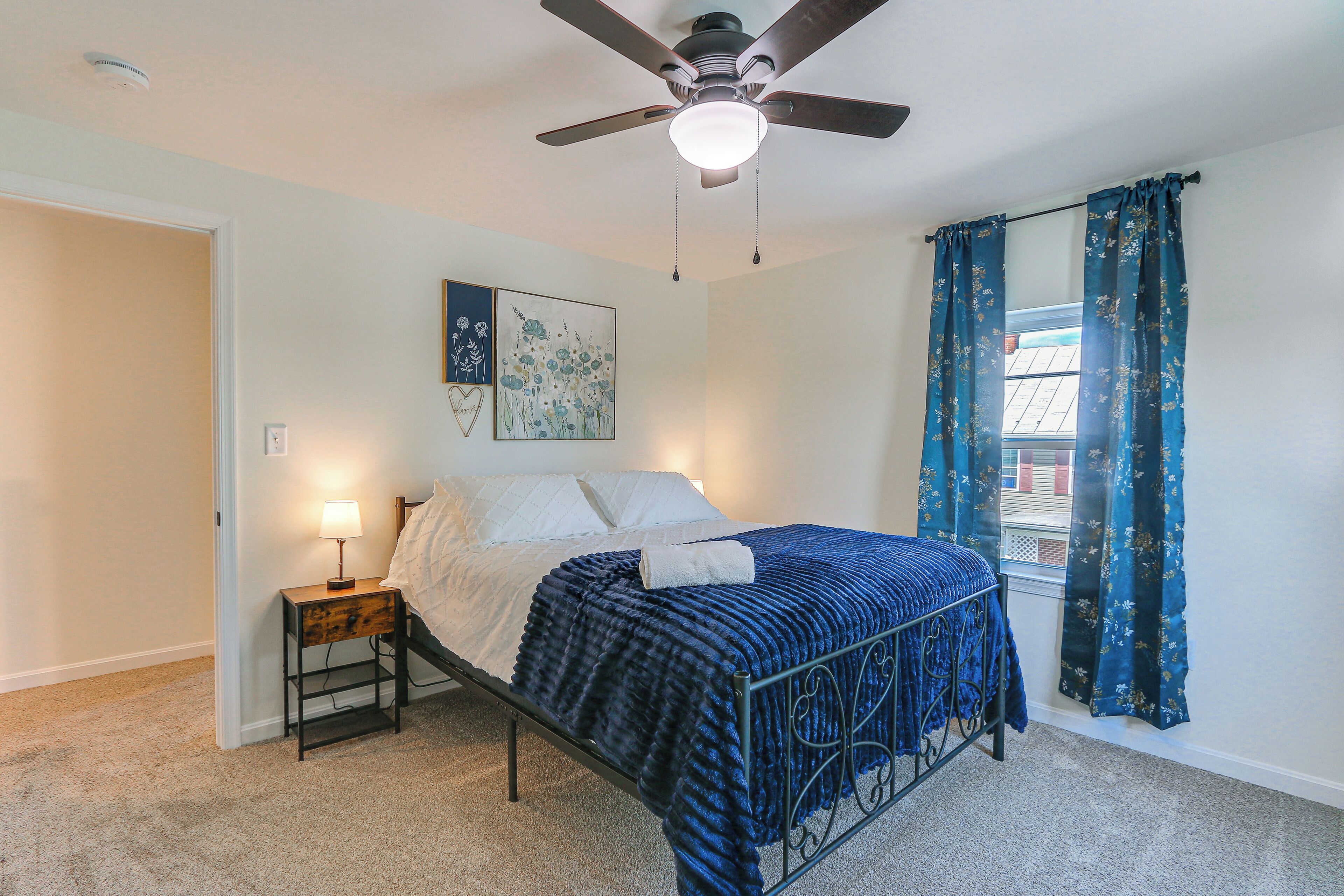 A bedroom with a bed, a nightstand, a ceiling fan, a window with curtains, and a wall-mounted light fixture.