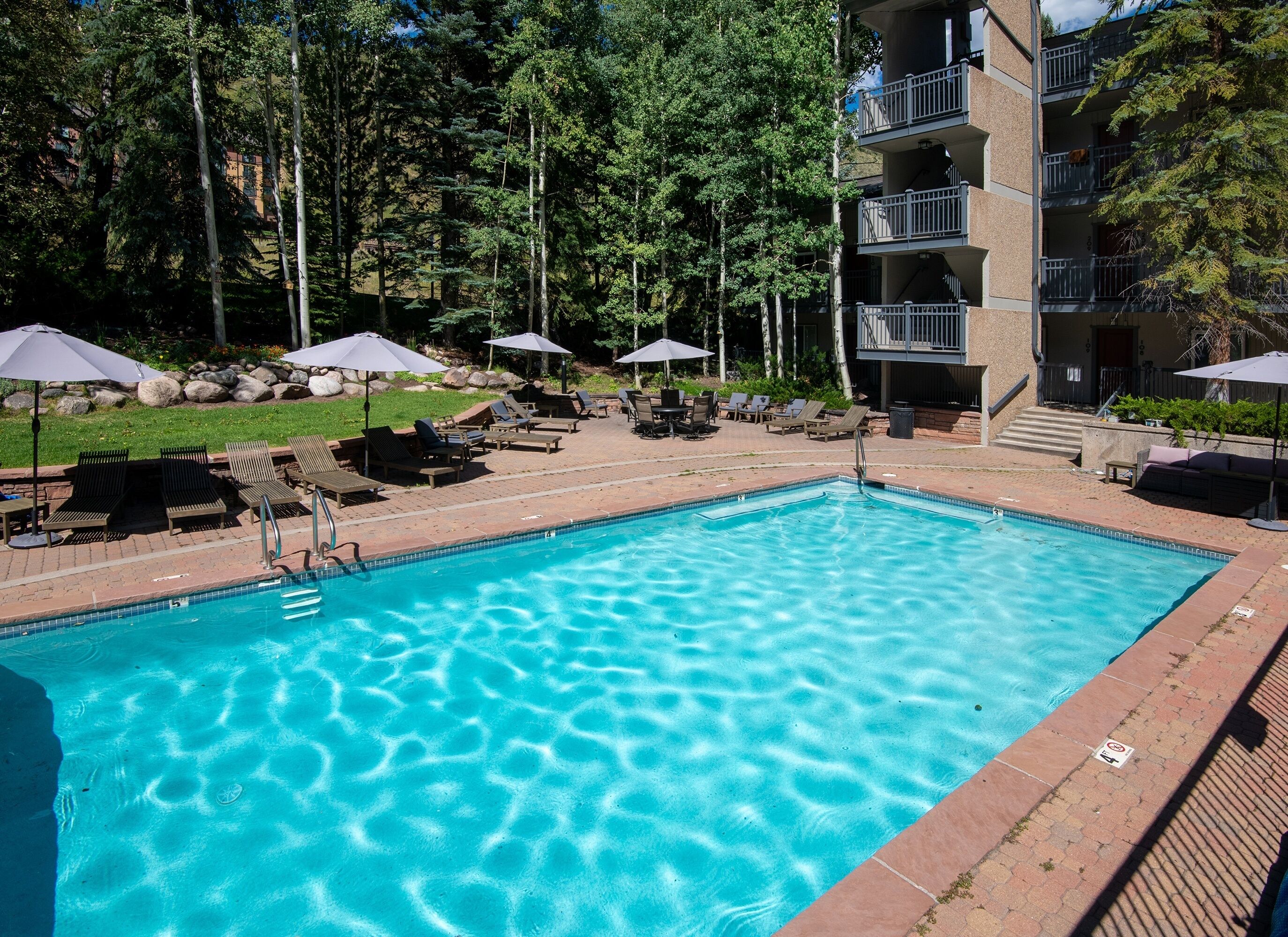 A rectangular swimming pool with clear blue water, surrounded by lounge chairs and umbrellas, set against a backdrop of trees and a multi-story building.