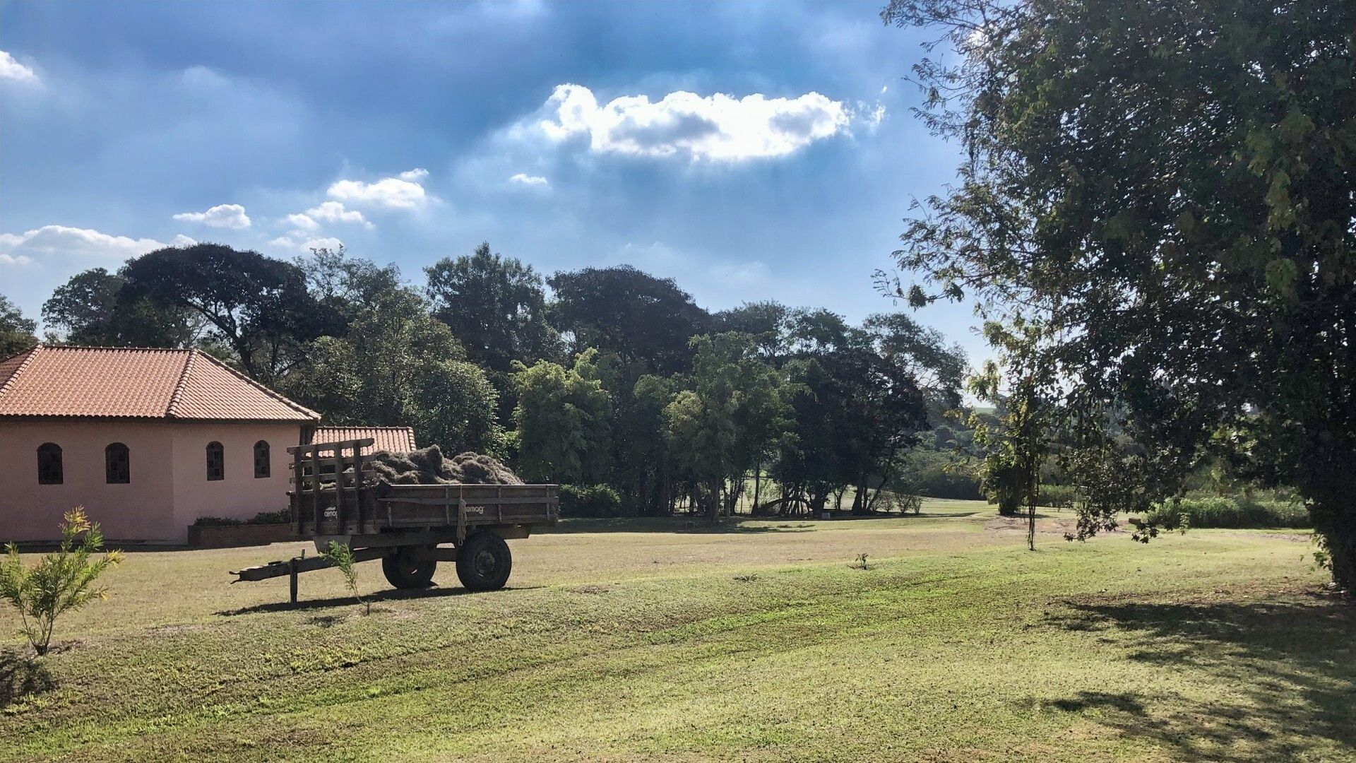 Ein Gebäude mit rotem Ziegeldach, ein Lastwagen mit Ladung und ein grasbewachsenes Feld mit Bäumen im Hintergrund.