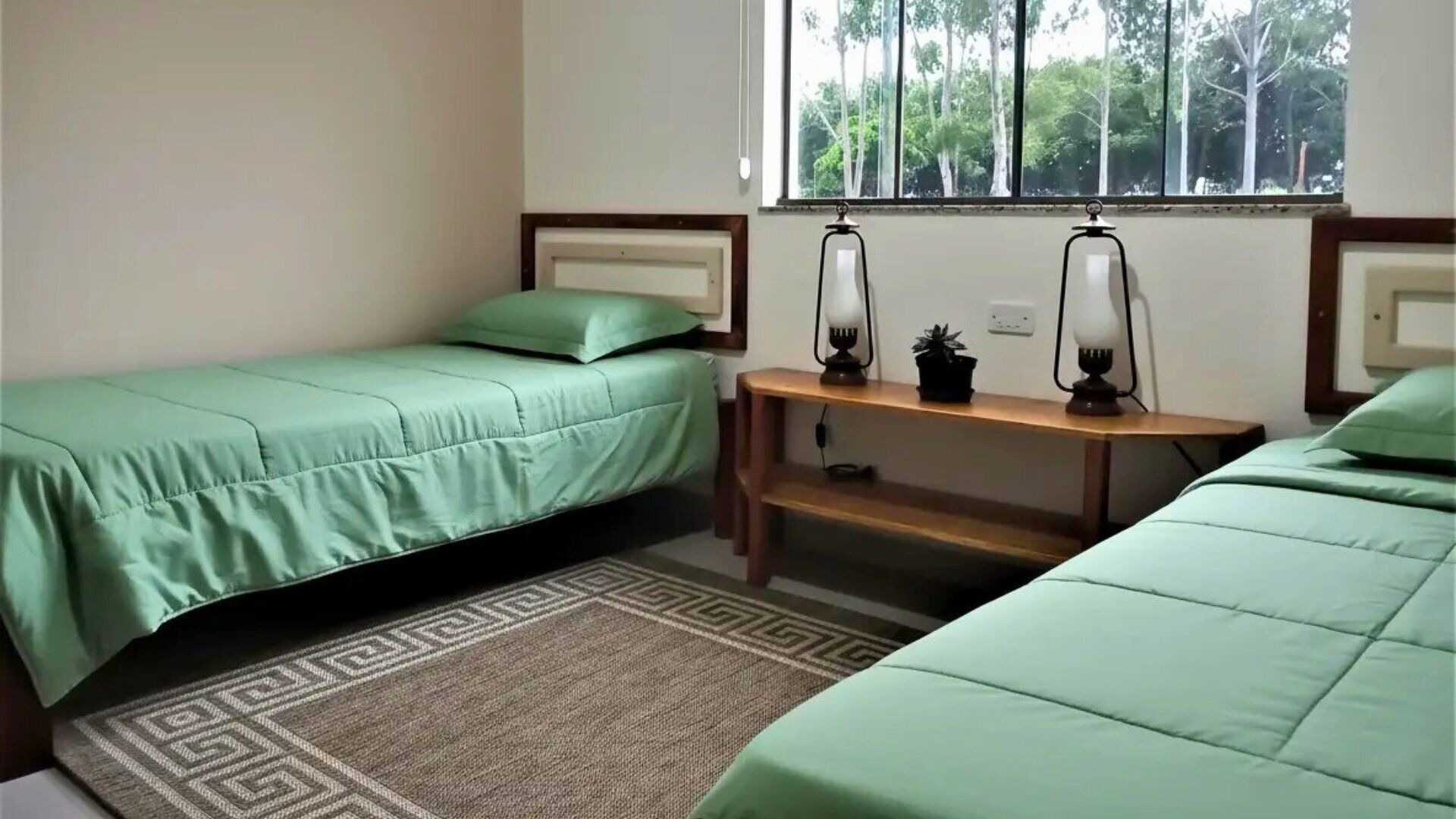 Une chambre avec deux lits simples, une petite table avec une lampe et une fenêtre donnant sur des arbres.