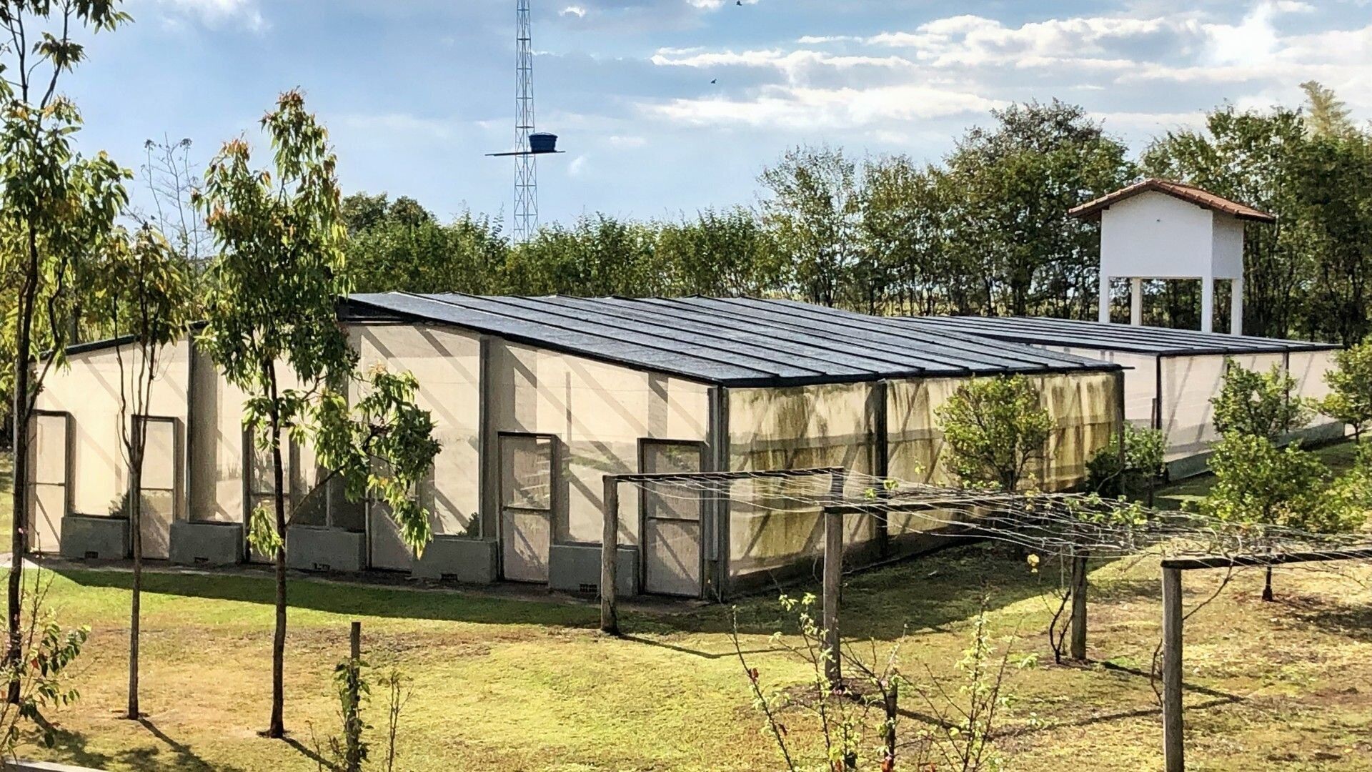 Een modern gebouw met een plat dak en grote ramen, omringd door bomen en een kleine witte constructie op de achtergrond.