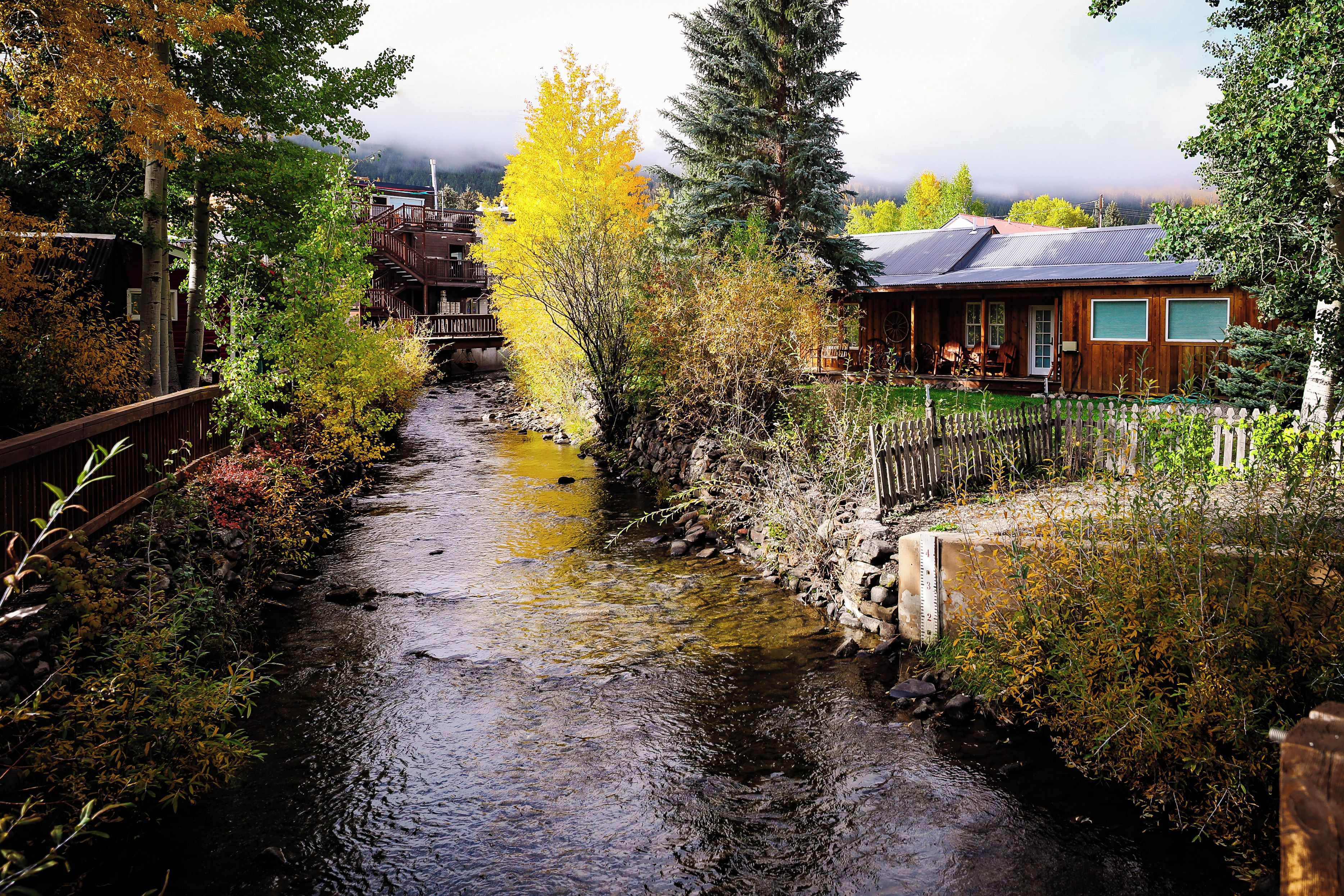 A narrow waterway flanked by trees and wooden structures, with a bridge in the background.